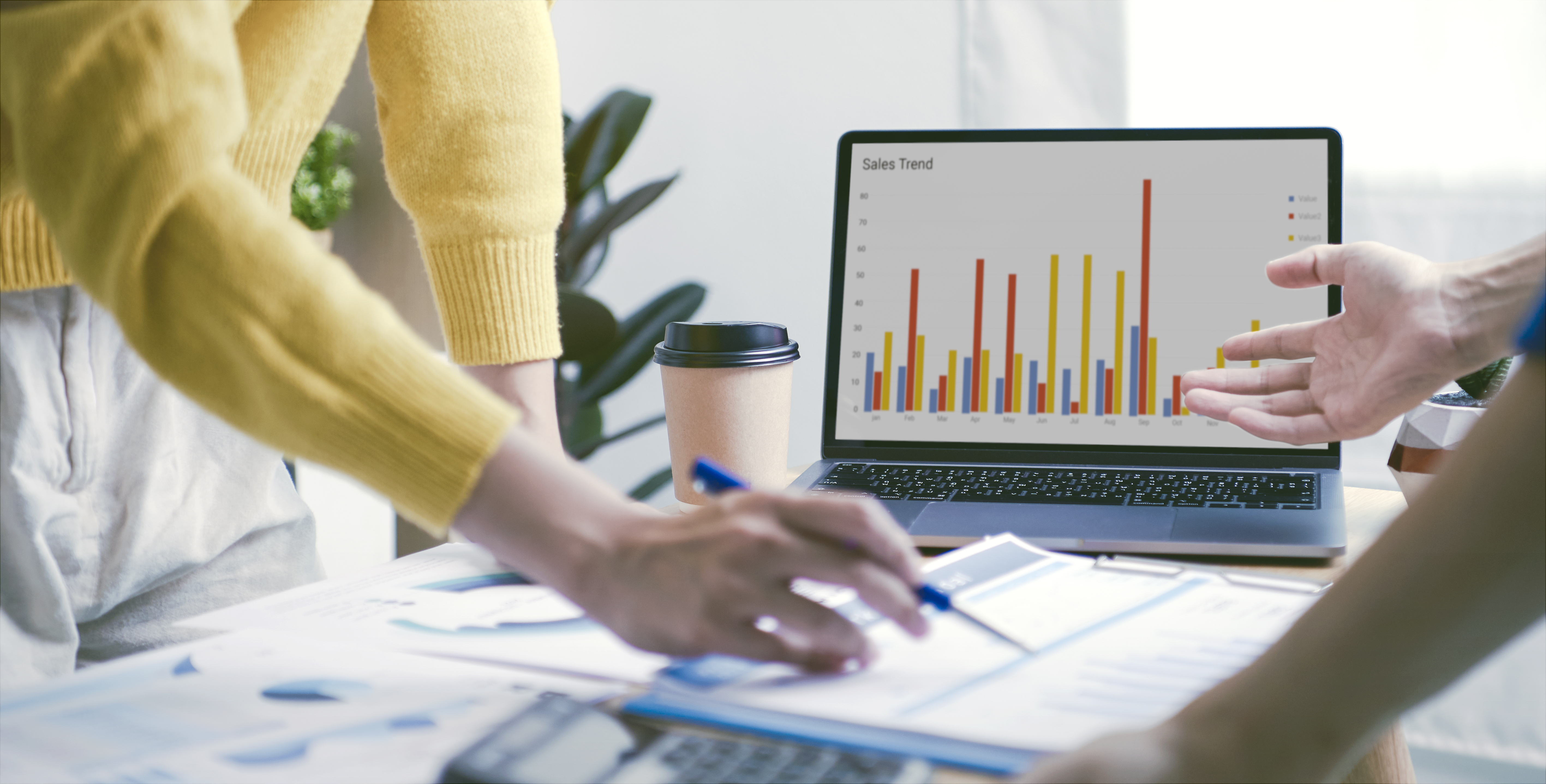 The image depicts a business meeting or discussion focused on analyzing sales trends. On the table, there are papers, a calculator, and a coffee cup, while a laptop screen displays a bar chart with different colors representing sales data over the months. One person is gesturing towards the screen, while another is taking notes, suggesting active data analysis and teamwork.